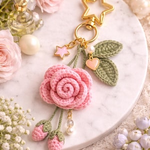 May include: A crochet keyring featuring a pink rose flower, two small pink buds with green stems, and two green crocheted leaves. The accessory includes a gold-toned star-shaped clasp, a small pink star charm, a heart charm, and white beads.