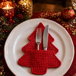 Pode incluir: Um suporte para talheres de crochê vermelho em forma de árvore de Natal, com um design de bolso que contém um garfo e uma faca de metal prateado, sobre um prato de cerâmica branca com borda festonada e acabamento liso e brilhante.
