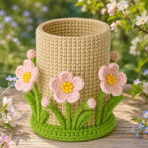May include: A cylindrical crochet pen holder in a light beige colour, featuring a vibrant green base and three dimensional light pink flowers with bright yellow centres. The flowers are attached to green stems and leaves around the exterior.