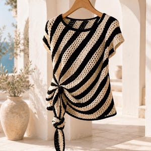 May include: A short-sleeved crochet top featuring a diagonal black and cream striped pattern. The garment has a round neckline and a prominent side knot detail that creates a draped effect, hanging down from the lower left side of the shirt.