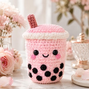 May include: A crocheted plush toy shaped like a pink boba tea cup. The cup features a white lid, a pink straw, black circular boba pearls, and a smiling face with black eyes and pink cheeks. The texture is soft and yarn-based throughout.