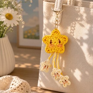 May include: A yellow crocheted star keychain with a smiling face and pink cheeks, featuring three smaller cream-colored crocheted stars dangling from the main star by thin yarn strands, attached to a silver metal keyring on a canvas tote bag.