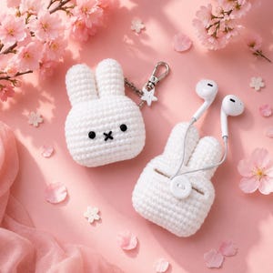 May include: Two white crocheted rabbit-shaped cases for wireless earbuds. One case features black embroidered eyes and a cross-stitched mouth with a silver metal keychain attachment. The other case holds white wired earbuds and has a small white button.