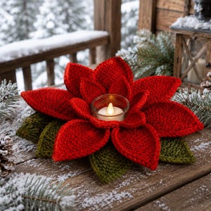 Può includere: Un portacandele a forma di stella di Natale all'uncinetto con petali rosso vivo e foglie verde scuro disposte a cerchio. Un bicchiere portacandela in vetro trasparente si trova al centro con una piccola candela bianca a batteria.