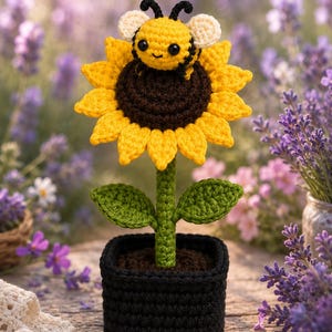 Puede incluir: Un girasol de ganchillo en una maceta cuadrada negra con una pequeña abeja amarilla y negra sobre el centro marrón oscuro. La flor tiene pétalos de color amarillo brillante, un tallo verde y dos hojas verdes hechas de hilo.