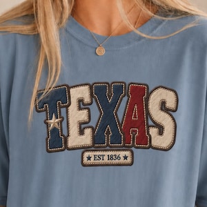 May include: A dusty blue short-sleeve t-shirt featuring the word TEXAS in large, block-style letters with a stitched border. The letters alternate between navy, cream, and red colours. Below the word is a small rectangular patch reading EST 1836.