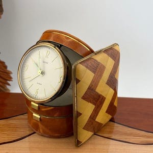 May include: A vintage travel alarm clock housed in a round, tan leather-covered case. The clock face is off-white with gold-tone markers and hands, featuring the word "IMAGE" and "ALARM" printed on it. The case has a hinged, geometric wood-inlay lid.