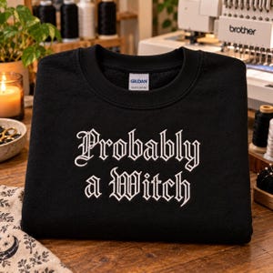 May include: A black crewneck sweatshirt featuring white embroidered text that reads "Probably a Witch" in a gothic-style font. The garment has a Gildan brand label on the inside collar and is made of a heavy blend fabric with long sleeves.