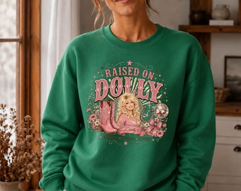 May include: A green crewneck sweatshirt featuring a graphic print with the text "RAISED ON DOLLY" in pink letters. The design includes an illustration of a blonde woman, pink cowboy boots, a disco ball, flowers, and a horseshoe in pink and green.
