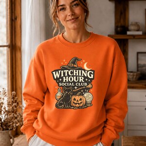 May include: An orange long-sleeve sweatshirt featuring a graphic print with the text "WITCHING HOUR SOCIAL CLUB". The design includes a black witch hat, a crescent moon, a black cat, a jack-o'-lantern, a potion bottle, and a broomstick.