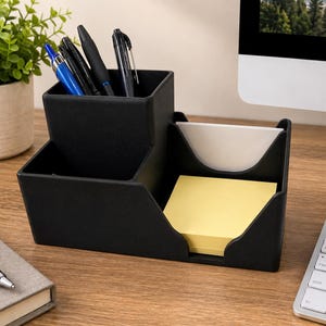May include: A black desk organizer featuring a tall square compartment holding four pens, a smaller front compartment, and a side section containing a stack of yellow sticky notes and a white notepad with a curved cutout for easy access.