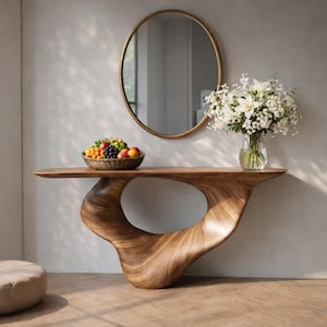 Op de afbeelding: Een moderne houten console met een uniek, organisch ontwerp. De tafel heeft een gebogen basis met een grote opening. Boven de tafel hangt een ronde gouden spiegel, waarop een gevlochten fruitmand en een vaas met witte bloemen staan.