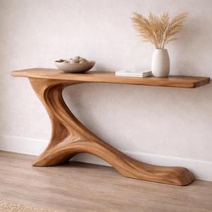 Può includere: Una consolle in legno dal design unico e organico. Il tavolo presenta una gamba curva e scultorea e un piano lungo e rettangolare. Una ciotola in ceramica con sfere decorative, un vaso con piante essiccate e un libro sono esposti sul tavolo.