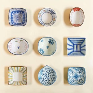 May include: A collection of nine small ceramic dishes arranged in a grid. The dishes feature various shapes including square, round, and oval, decorated with blue, green, and red hand-painted patterns such as stripes, floral motifs, and geometric designs.