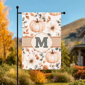 May include: A rectangular garden flag featuring a white background with a pattern of light orange pumpkins, white flowers, and autumn leaves. A horizontal tan band crosses the center with a scalloped circular frame containing the letter M.