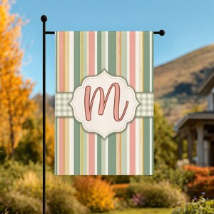 May include: A rectangular garden flag featuring a vertical striped pattern in shades of sage green, soft pink, and cream. A central cream-colored scalloped label displays a large, stylized letter M in pink, set against a horizontal gingham band.