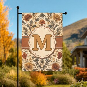 May include: A rectangular garden flag featuring a cream background with a repeating pattern of orange and yellow chrysanthemums and dark green foliage. A horizontal brown band crosses the center, overlaid with a scalloped cream frame containing a large, bold, tan letter M.