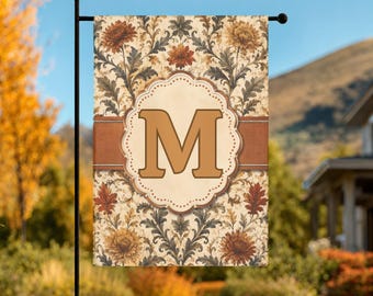Könnte beinhalten: Eine rechteckige Gartenflagge mit cremefarbenem Hintergrund und einem sich wiederholenden Muster aus orangefarbenen und gelben Chrysanthemen sowie dunkelgrünem Blattwerk. Ein horizontales braunes Band verläuft durch die Mitte, überlagert von einem cremefarbenen, gewellten Rahmen mit einem großen, fetten, beigen Buchstaben M.