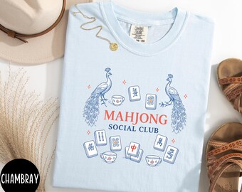 May include: A light blue t-shirt featuring a graphic print of two peacocks, several mahjong tiles, and three teacups. The text "MAHJONG SOCIAL CLUB" is printed in the center in red and blue. The shirt has a crew neckline and a tag.