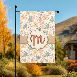 May include: A rectangular garden flag featuring a repeating pattern of autumn leaves and berries in shades of orange, yellow, and sage green. A horizontal burlap-textured band crosses the center with a central cream-colored scalloped label displaying the letter M.