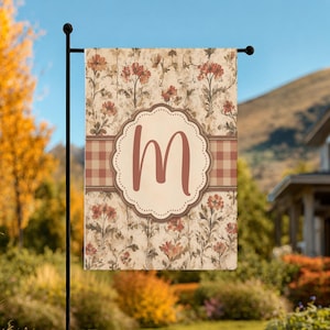 May include: A rectangular garden flag featuring a vintage-style floral pattern in muted orange and brown tones. A horizontal brown and white gingham stripe crosses the center, overlaid with a scalloped cream-colored circle containing the letter M.
