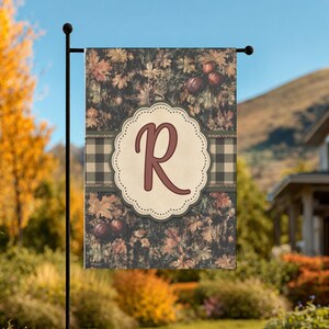May include: A rectangular garden flag featuring a dark autumn leaf pattern with small pumpkins and a horizontal black and white buffalo plaid stripe. A central cream-colored scalloped emblem displays a large, dark red letter R in script font.