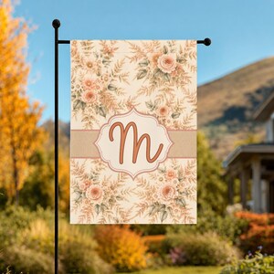 May include: A rectangular garden flag featuring a cream-colored background with a repeating pattern of soft peach-toned flowers and fern-like foliage. A horizontal band crosses the center with a central decorative frame containing the letter M.