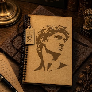 May include: A spiral-bound notebook with a tan cover featuring a dark brown silhouette of Michelangelo's David. A small rectangular tag hangs from the black wire binding, printed with the text ART COLLECTION DAVID MICHELANGELO and a small portrait.