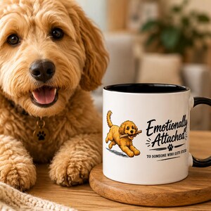 May include: A white mug with a black handle and rim, displaying a cartoon dog with a stick and the text "Emotionally Attached to Someone Who Eats Sticks." A golden doodle dog is in the background.