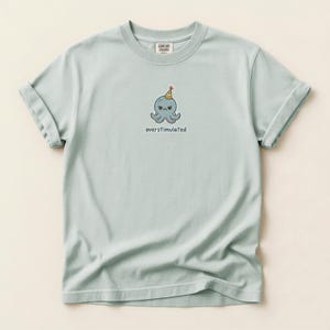 以下が含まれることがあります： 明るいセージグリーンの半袖Tシャツ。カラフルなパーティーハットをかぶった不機嫌そうな青いタコの小さなグラフィックが描かれています。タコの下には、シンプルな暗い小文字のサンセリフフォントで「overstimulated」という文字がプリントされています。