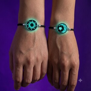 May include: Two black cord bracelets featuring circular silver-toned charms. One charm displays a glowing cyan sun design, while the other charm features a glowing cyan crescent moon and star design, both set against a dark background.