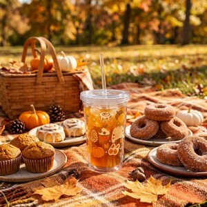 Puede incluir: Un vaso de plástico transparente con pajita, lleno de líquido ámbar y decorado con iconos de temática otoñal que incluyen calabazas, rollos de canela y piñas, colocado entre platos de muffins, rosquillas y rollos de canela.