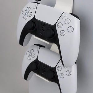 Puede incluir: Un soporte doble de plástico blanco para mandos, diseñado para colgarse en el lateral de una consola de juegos. El soporte sostiene dos mandos inalámbricos blancos y negros, cada uno con botones direccionales estándar, joysticks y el logotipo de PlayStation.