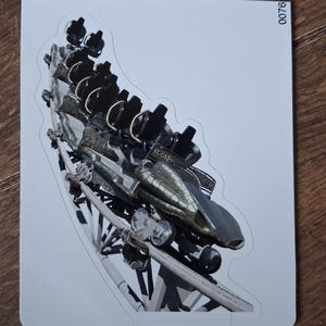 May include: A die-cut sticker featuring a roller coaster train on a white track. The train is black with a metallic gold front section and the word ICON printed on the side. The sticker has a white border and the number 0076 printed in the top right corner.