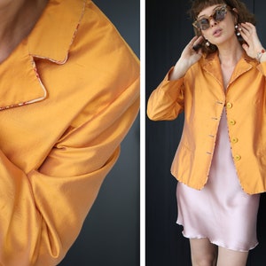 May include: A mustard yellow blazer with a contrasting trim around the collar and edges. The blazer has a button closure and is made of a silky fabric.