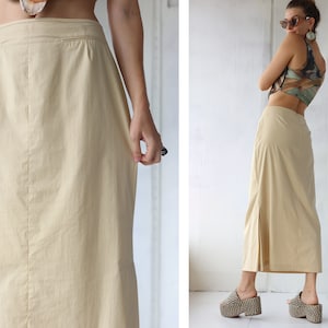 May include: A light beige maxi skirt with a straight silhouette and a side slit. The skirt is made of a lightweight fabric and has a simple design.