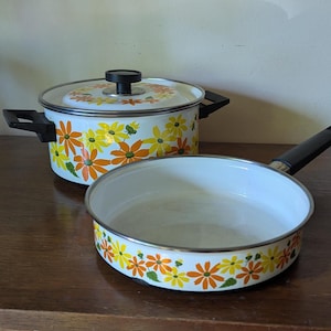May include: A vintage white enamel cooking pot with a lid and a matching frying pan. Both pieces are decorated with a floral pattern in orange, yellow, and green. The pot has black handles and a black knob.