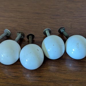 May include: Six vintage white ceramic cabinet knobs with metal screws, arranged on a wooden surface. The knobs are round and have a glossy finish, showing signs of age. Ideal for furniture restoration or DIY projects.