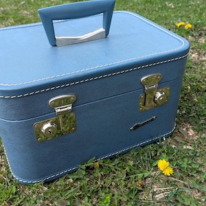 May include: A vintage blue suitcase with a handle and brass-colored latches. The case has white stitching around the edges and sits on a grassy surface. The handle is a matching blue color with a silver-colored metal accent.