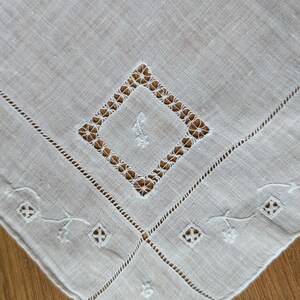 May include: Off-white linen handkerchief with a central diamond-shaped openwork design. The corners feature embroidered floral and geometric patterns. The edges are finished with a delicate stitched border, showcasing a vintage aesthetic.