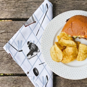May include: A white cloth napkin featuring a light blue diamond grid pattern with hockey-themed illustrations including black hockey skates, a black hockey puck, and hockey sticks, positioned next to a white plate with a burger and potato chips.