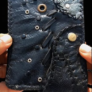 Op de afbeelding: Een zwarte lederen portemonnee met een textuur die bestaat uit een mix van gladde en generfde afwerkingen. Het ontwerp bevat decoratieve metalen ringen, zwarte studs, lederen veterdetails en een enkele lichtgekleurde ronde knoopsluiting.