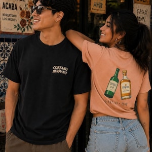 May include: A black t-shirt with the text COREANO HERMANO on the chest and a peach-colored t-shirt featuring a graphic of a green soju bottle and a gold tequila bottle with the text SALUD and Korean characters printed on the back.