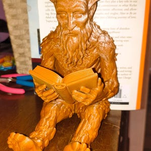 Op de afbeelding: Een bruin, driedimensionaal geprint beeldje van een zittend boomachtig wezen met een menselijk gezicht, baard en takken als haar. Het figuur houdt een open boek in de handen, met de benen naar voren gestrekt en de voeten zichtbaar aan de basis.