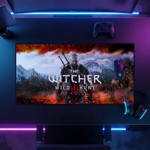 May include: A rectangular desk mat featuring a graphic print of The Witcher 3 Wild Hunt with a central character figure, snowy mountains, and orange embers. The mat displays the game title in white text against a dark, atmospheric background.
