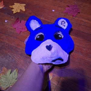May include: A blue bear-shaped mask with a light purple muzzle, a black heart-shaped nose, and black mesh eye coverings. The mask features rounded ears and a small white patch on the left ear with a black cloud and raindrop graphic design.
