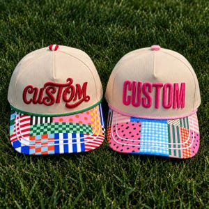 May include: Two beige baseball caps with structured crowns and top buttons. The left cap features red cursive embroidery reading custom and a multicolored patchwork brim. The right cap has hot pink block letter embroidery reading CUSTOM and a patterned brim.