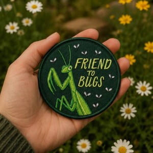 May include: A circular embroidered patch featuring a bright green praying mantis on a dark green background. The text "FRIEND TO BUGS" is stitched in light green letters, accompanied by small white embroidered insects scattered around the design.