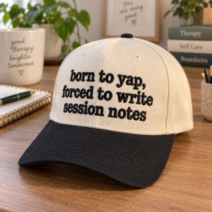 May include: A baseball cap with a cream-colored crown and a black brim. The front of the crown features black embroidered text that reads, "born to yap, forced to write session notes," centered in a clean, sans-serif font with a small black button.