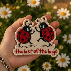 May include: An embroidered patch featuring two cartoon ladybugs with red wings and black spots, waving at each other. The patch has a cream-colored background and includes the red embroidered text "the last of the bugs" at the bottom.